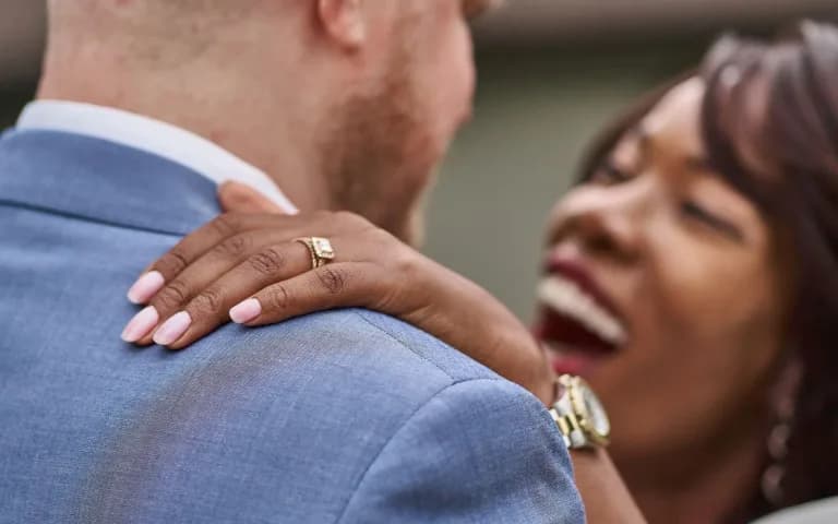 Close de um casal se abraçando, a mão dela com aliança apoiada no ombro dele, sorriso espontâneo.