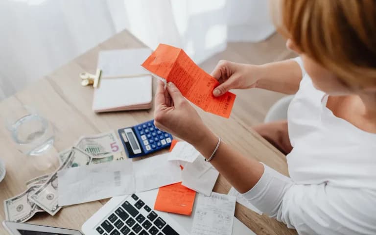 Close view of a woman sorting receipts and using a calculator at a home desk.