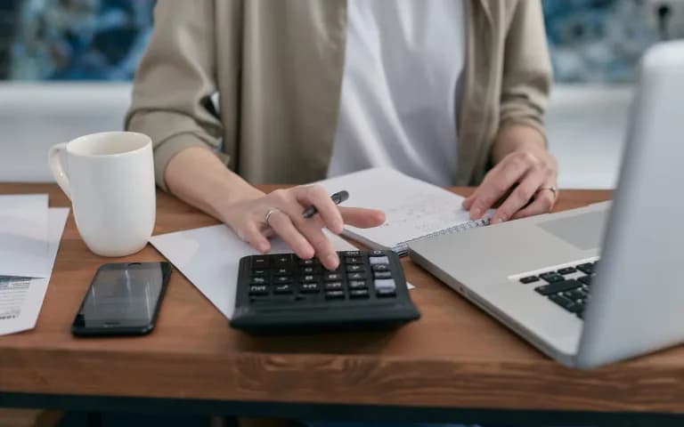 Persona usando una calculadora sobre una mesa de madera, con laptop, libreta y una taza de café al lado.