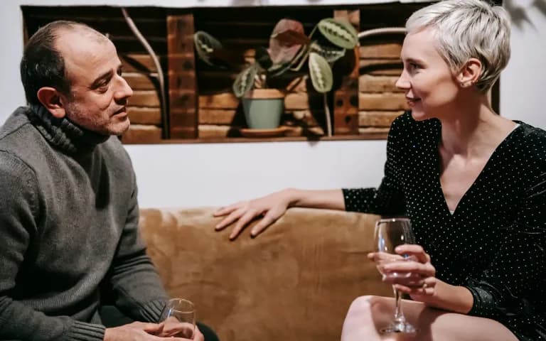 Casal sentado no sofá em casa, conversando com atenção um com o outro, taças na mão, clima calmo e acolhedor.