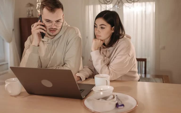 Casal de moletom concentrado, usando notebook e celular juntos em casa.