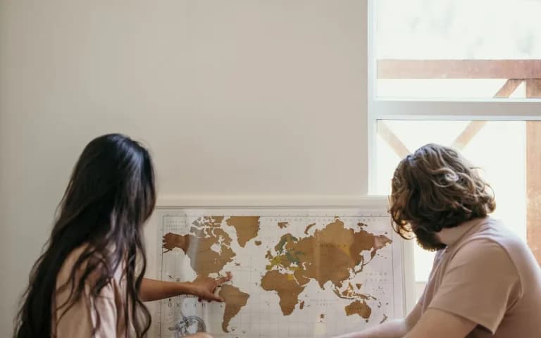 Casal sentado no chão de casa, apontando juntos para um mapa-múndi pendurado na parede, planejando a próxima viagem.