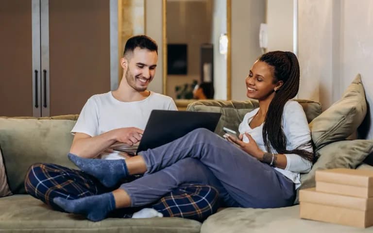 Casal de pijama sorrindo no sofá, usando notebook e celular juntos, com caixas de compras ao lado.