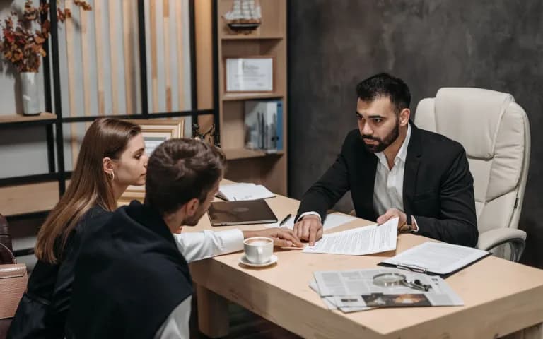 Advogado de terno conversando com um casal de clientes em uma mesa de escritório.