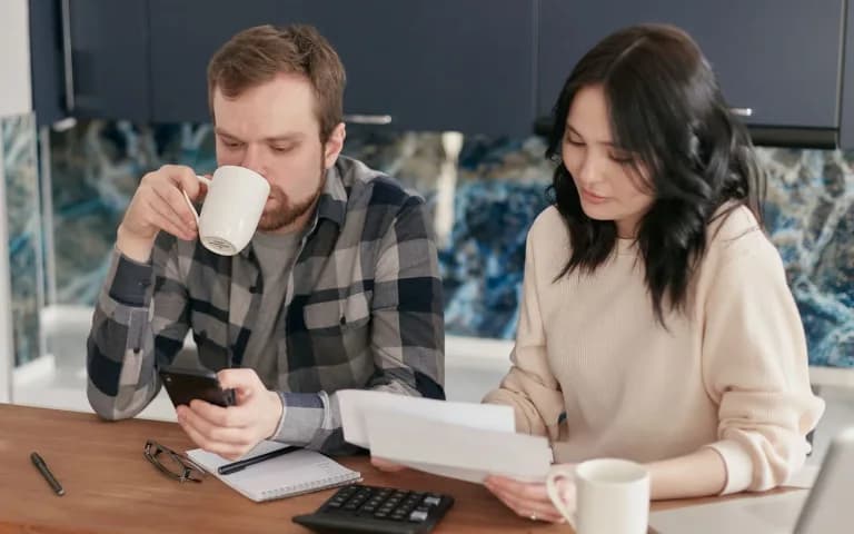 Casal na bancada da cozinha, ele tomando café e olhando o celular, ela lendo um extrato impresso, com calculadora e caderno sobre a mesa.