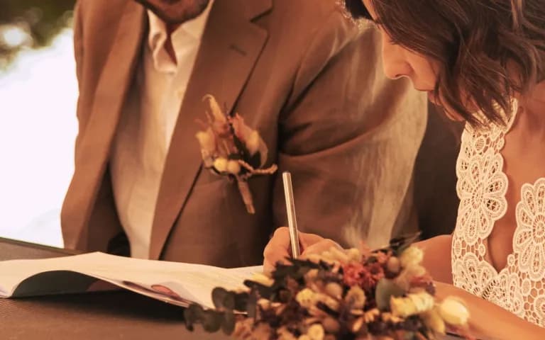 Noivo observando enquanto a noiva assina o documento oficial do casamento civil, ao ar livre.
